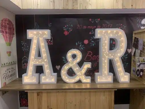 Imagem da promoção do Pacote Letras Iluminadas, com as iniciais A&R e os dois presentes em miniatura sobre uma mesa, em cenário de casamento.