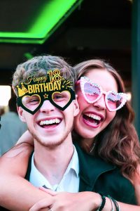 Dois jovens sorridentes usam óculos divertidos de feliz aniversário numa festa com decoração de aniversário 18 anos.