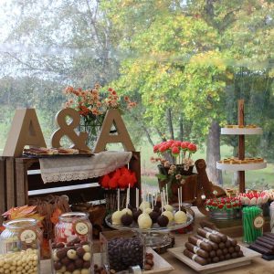 Letras gigantes luminosas com as iniciais de casamento J e M a brilhar ao lado de uma mesa de doces decorada com flores.