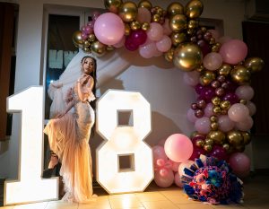Jovem com vestido de gala posa junto a números gigantes iluminados numa decoração de aniversário 18 anos com balões cor-de-rosa e dourados.
