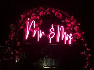 Letreiro de neon rosa brilhante com a inscrição Mr & Mrs integrado num arco decorativo com flores escuras durante uma festa de casamento à noite.