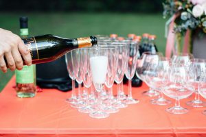 Mão a servir espumante ou prosecco numa taça de vidro flute sobre uma mesa decorada num casamento ao ar livre.