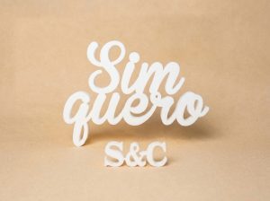 Letras caligráficas brancas de madeira com a frase Sim quero sobre um fundo bege numa festa de noivado original.