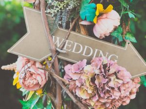 Placa decorativa de madeira em forma de seta com a palavra WEDDING e flores brancas como ideias para festa de noivado.