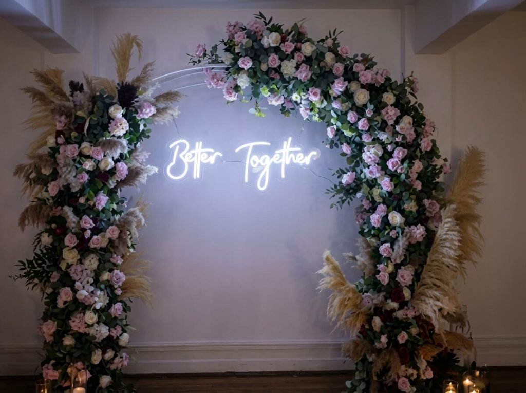 Cenário para festas com arco redondo de flores, pampas secas e letreiro néon luminoso Better Together.