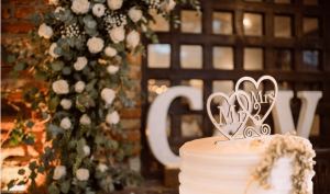 Cenário de casamento com letras XXL iluminadas por trás da mesa do bolo com arranjo floral rústico.