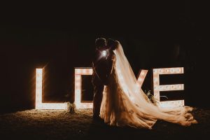 Noivos a beijarem-se à noite em frente a letras iluminadas LOVE num casamento ao ar livre.