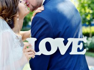 Primeiro plano de um casal de noivos a beijar-se enquanto a noiva segura umas letras de madeira branca com a palavra LOVE.