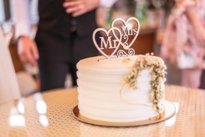 Bolo de casamento texturizado com topo de madeira personalizado de corações e iniciais Mr e Mrs com iniciais desfocadas ao fundo.